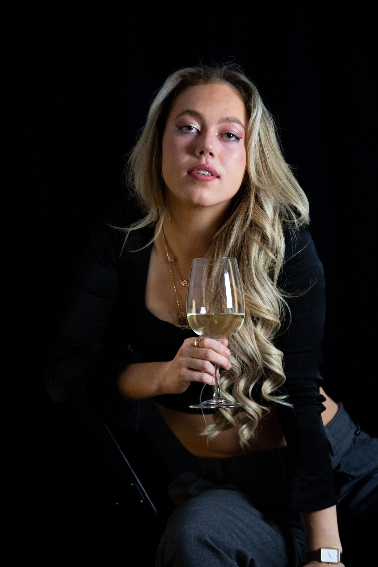 Stylish woman with long blonde hair holding a white wine glass against a black background.