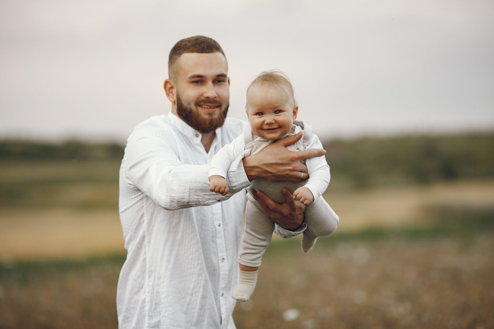 Smiling bearded father dressed in white holds his happy baby outdoors on a sunny day.