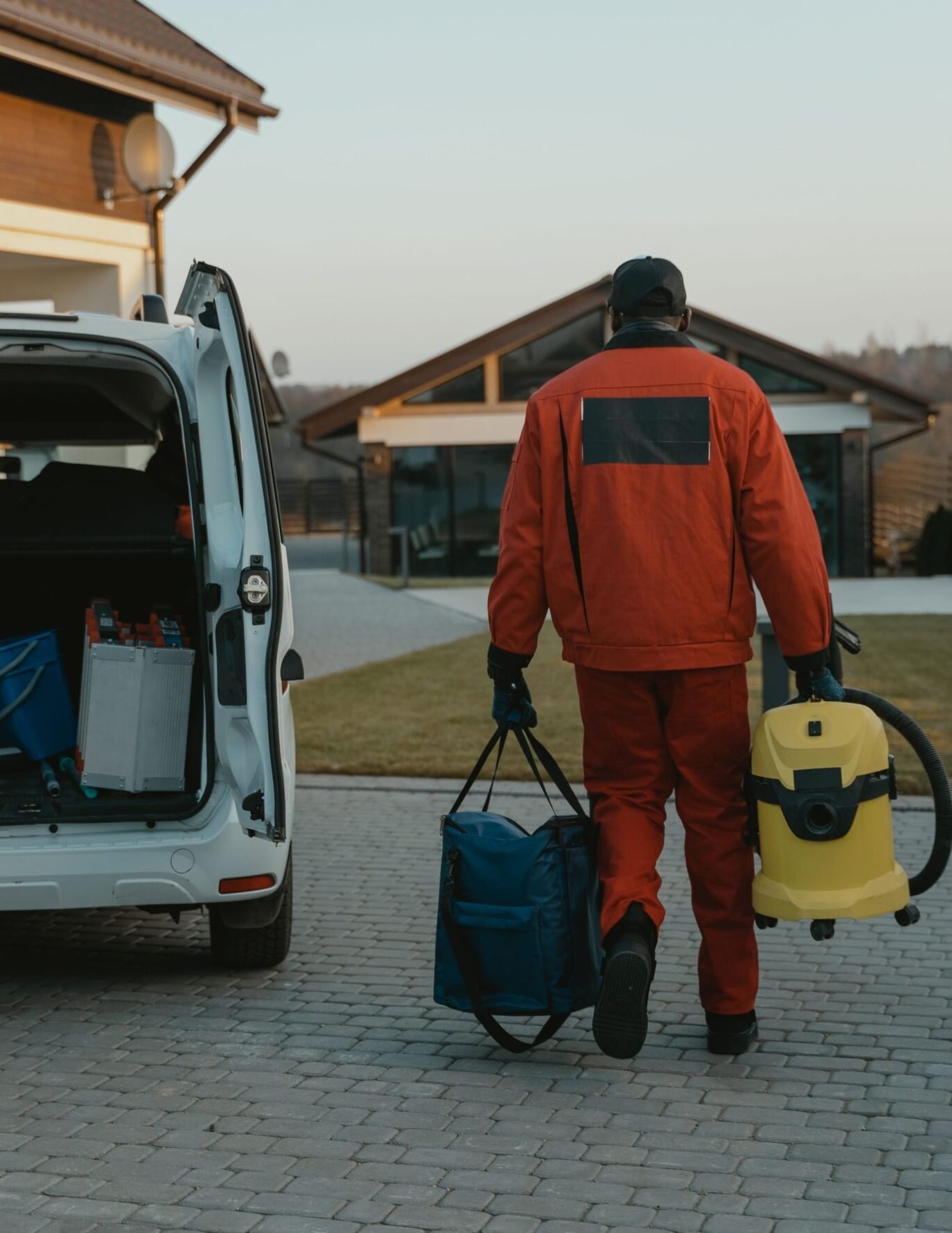 A worker in red work clothes carries a vacuum cleaner and bag near a van during the day.